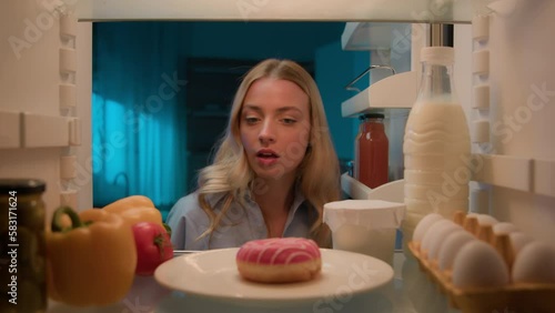 Point of view POV from inside refrigerator hungry woman girl at night kitchen open fridge looking around eating bite sweet donut unhealthy nutrition diet failure weight control obesity food addiction photo