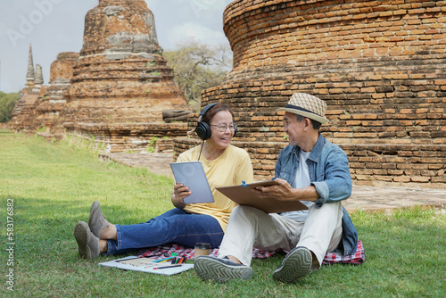 happy senior couple enjoy outdoors leisure time in Ayutthaya Historical Park,Thailand. the old man drawing picture and older adult woman playing on a digital tablet to listen music              