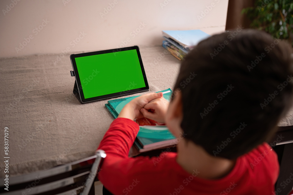 Rear view: smart student sitting a table a watching online lesson on a digital tablet with empty green chroma key screen with copy space for advertising text or mobile application. Distance education