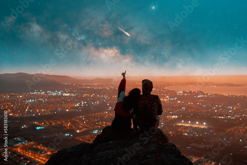 silhouette rear view of a heterosexual couple sitting on top of a mountain watching the starry sky with milky way and meteorite over the city