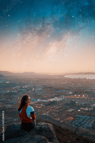 rear view young caucasian woman sitting on the top of the hill looking at the horizon relaxed with city and milky way in the background