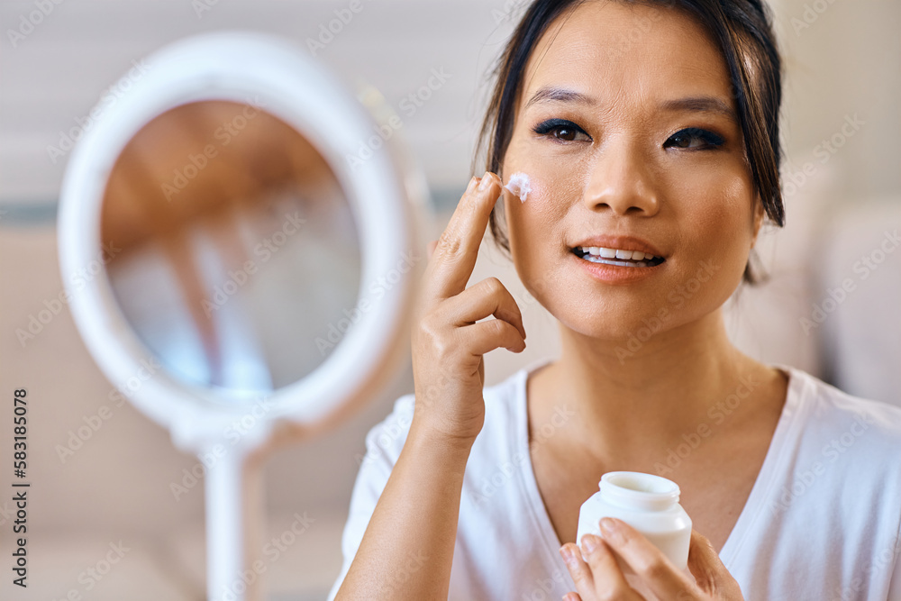 Asian woman applying face cream while looking herself in mirror. Asian woman applying face cream while looking herself in mirror.