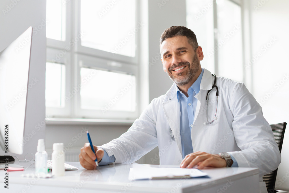 Happy middle aged male doctor in white medical uniform sitting at desk in hospital and smiling at camera, copy space Happy middle aged male doctor in white medical uniform sitting at desk in hospital and smiling at camera, copy space