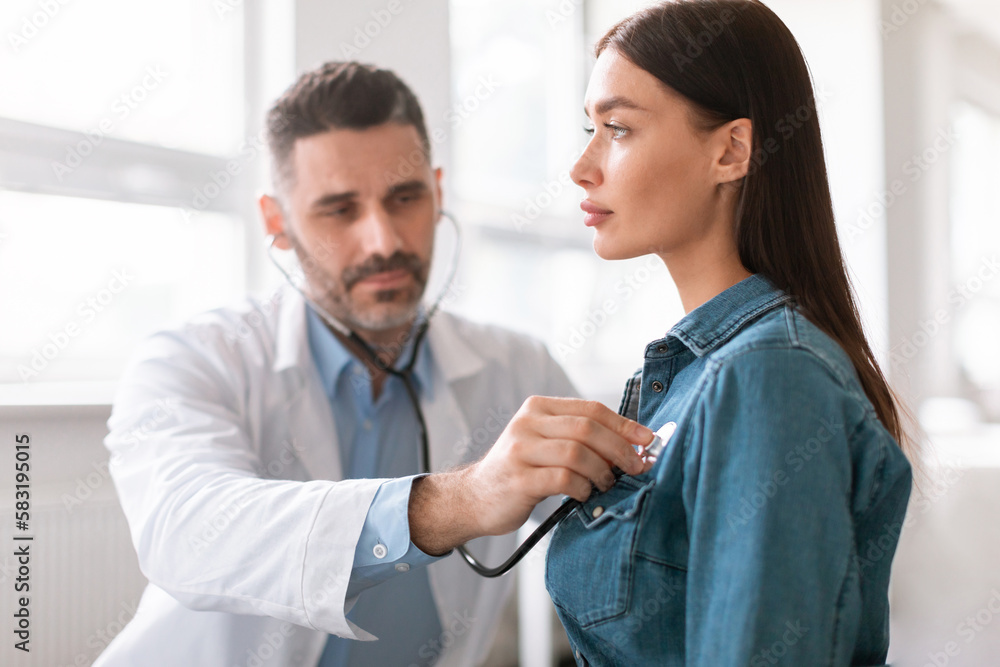 Lady coming to clinic for checkup, male doctor using stethoscope, listening to patient's breath or heartbeat Lady coming to clinic for checkup, male doctor using stethoscope, listening to patient's breath or heartbeat