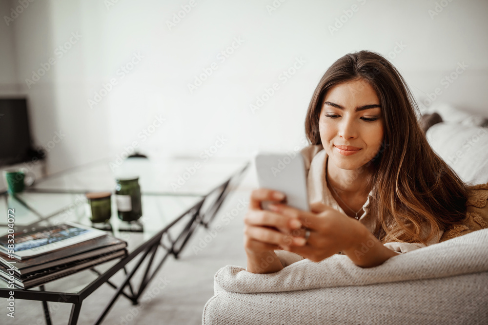 Cheerful millennial mixed race female look at phone, read message, chatting, enjoy free time, lies at sofa Cheerful millennial mixed race female look at phone, read message, chatting, enjoy free time, lies at sofa