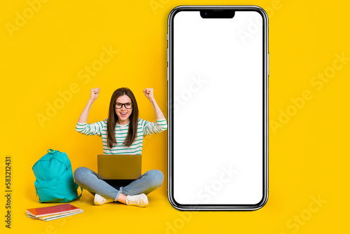 Photo of cheerful laughing girl raise fists in success pass college test sit next to phone promo isolated on yellow color background