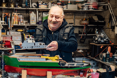Senior man, pensioner making ship model building handcraft on table with different materials in his garage. Retired elderly man boat modeling hobby. Active Retirement lifestyle. Selective focus.
