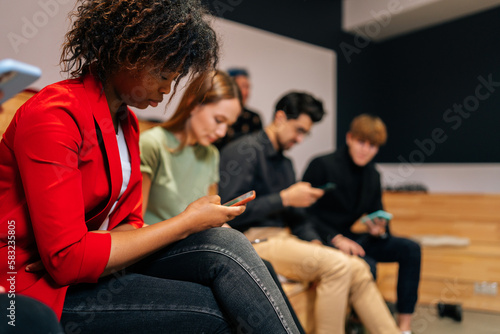 Close-up of multiethnic young men and women sitting along wall, looking at screens of smartphones, reading news, text messaging, chatting with friends, connecting to free wifi, waiting something.