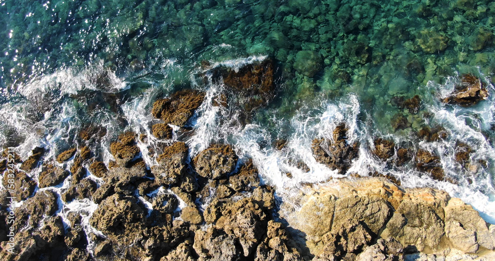 Obraz premium Aerial top view over the island's rocky shore and clear sea with varying shades of blue and light foamy waves in warm summer weather.