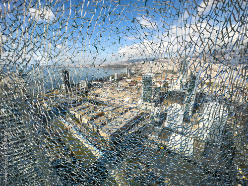 The blue sky, clouds, sea and city are visible through broken glass from the skyscraper, top angle view. Unique crack window with scenic background. Safety danger security concept. Filigree pattern