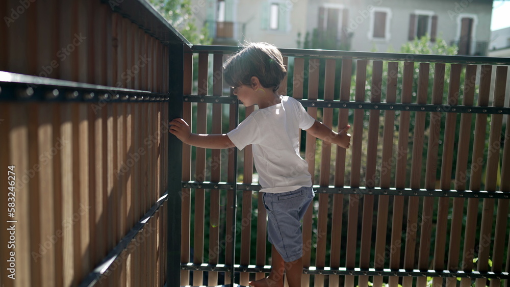 One playful little boy standing at apartment balcony in sunlight. Child holding on terrace fence with lens flare. Kid looking outside through wooden porch One playful little boy standing at apartment balcony in sunlight. Child holding on terrace fence with lens flare. Kid looking outside through wooden porch