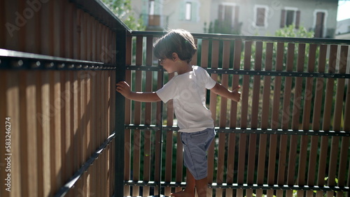 One playful little boy standing at apartment balcony in sunlight. Child holding on terrace fence with lens flare. Kid looking outside through wooden porch