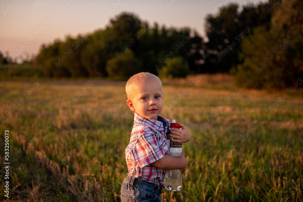 Little boy in plaid shirt holds bottle of water in hands, in mowing field of wheat in rays of sunset Little boy in plaid shirt holds bottle of water in hands, in mowing field of wheat in rays of sunset