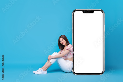 Photo of pretty positive lady sit big telephone board empty space demonstrates modern gadget shop sale isolated on blue color background © deagreez