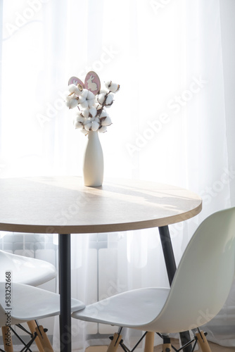A bouquet of cotton twigs in a vase on a round table with an Easter decor of eggs and an Easter bunny in the light from the window in a minimalist interior.