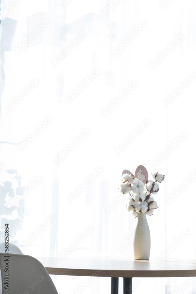 A bouquet of cotton twigs in a vase on a round table with an Easter decor of eggs and an Easter bunny in the light from the window in a minimalist interior. A bouquet of cotton twigs in a vase on a round table with an Easter decor of eggs and an Easter bunny in the light from the window in a minimalist interior.