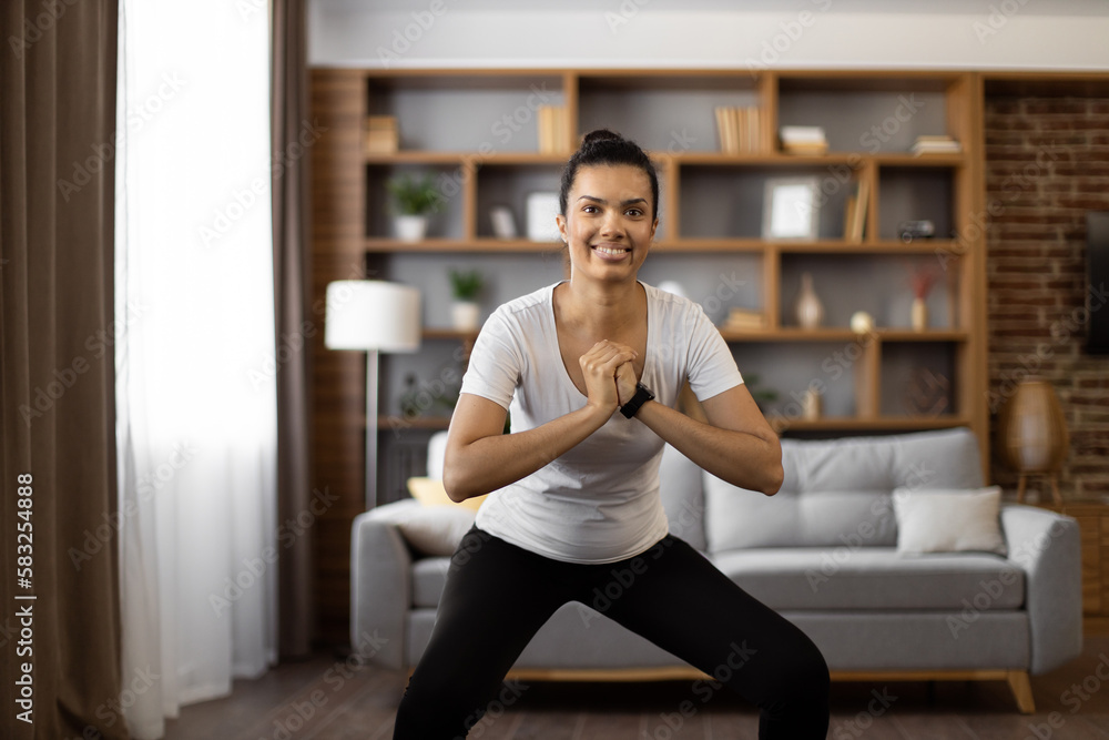 Fitness sport woman dressed in active outfit squatting while exercising alone at living room. Multiracial young female training regular at home for staying fit and healthy.