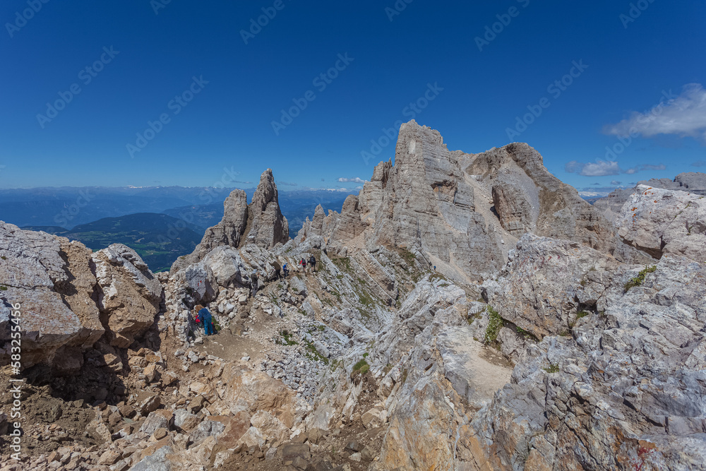 Trekkers along path in a awesome summer dolomite rocky scenario in the Latemar Massif, UNESCO world heritage site. The main pinnacle is named Torre di Pisa. Trentino-Alto Adige, Italy, Europe