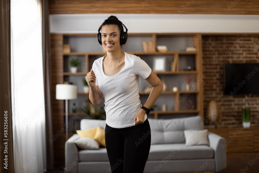 Active multiracial woman with dark hair enjoying music in wireless headphones while training at living room. Fitness young lady in sport clothes using modern gadget for workout.