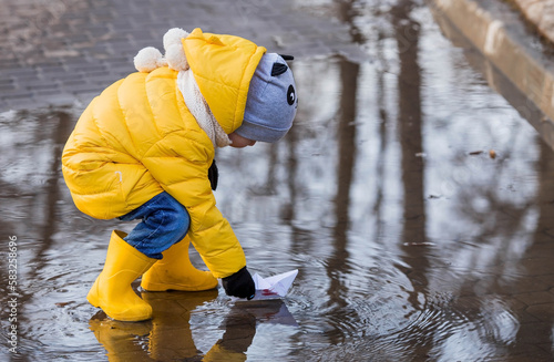 A small child in yellow rubber boots and a jacket runs through puddles, has fun, plays and launches paper boats. Spring break photo. It\'s springtime.