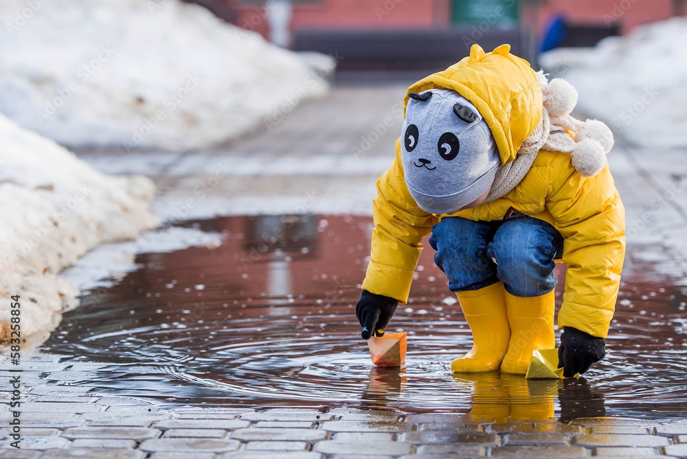 A small child in yellow rubber boots and a jacket runs through puddles, has fun, plays and launches paper boats. Spring break photo. It's springtime.