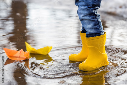 It's springtime. A small child in yellow rubber boots jumps through puddles, plays and launches paper boats on the water.. Photo of spring and autumn holidays. The concept of spring.