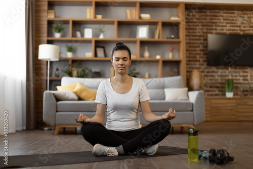 Peaceful interracial woman meditating in lotus pose with closed eyes while sitting on yoga mat. Young brunette in sportswear practicing breathing exercise at home.