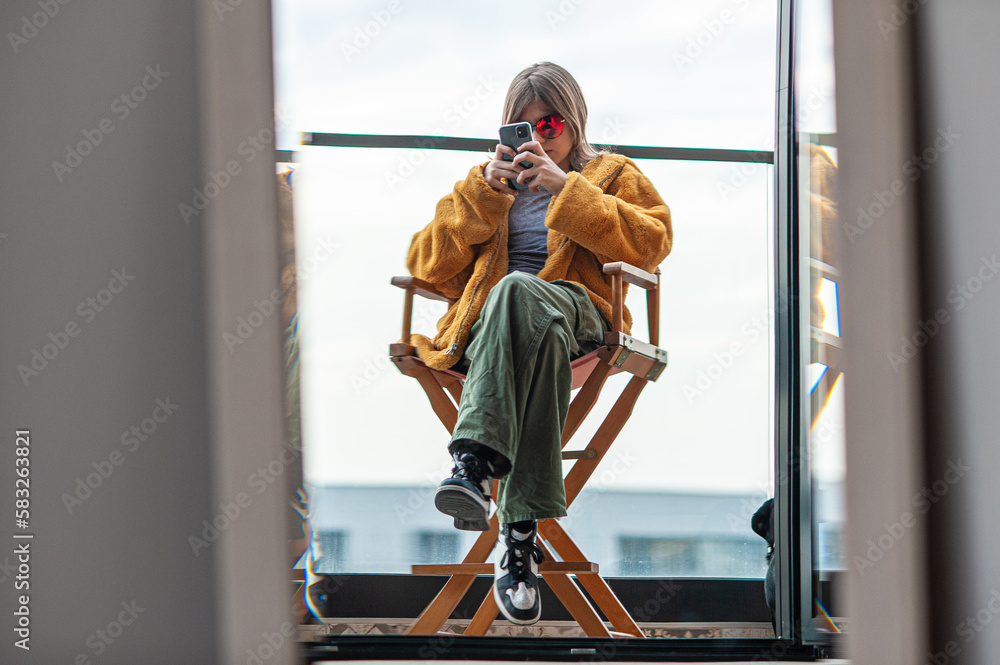 Beautiful young girl in a yellow fur coat is sitting in a director's chair on the terrace of a modern building. Using smartphone, social media influencer, taking picture, selfie, posing, city in backg Beautiful young girl in a yellow fur coat is sitting in a director's chair on the terrace of a modern building. Using smartphone, social media influencer, taking picture, selfie, posing, city in backg