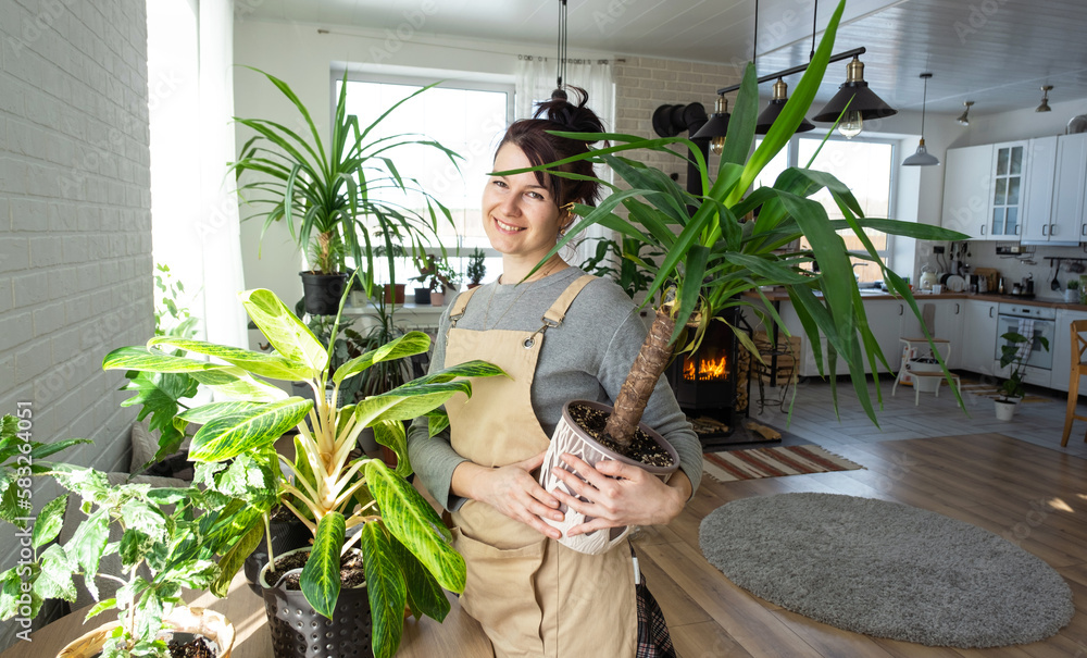 A happy woman in a green house with a potted plant in her hands smiles, takes care of a flower. The interior of a cozy eco-friendly house, a fireplace stove, a hobby for growing and breeding homeplant A happy woman in a green house with a potted plant in her hands smiles, takes care of a flower. The interior of a cozy eco-friendly house, a fireplace stove, a hobby for growing and breeding homeplant