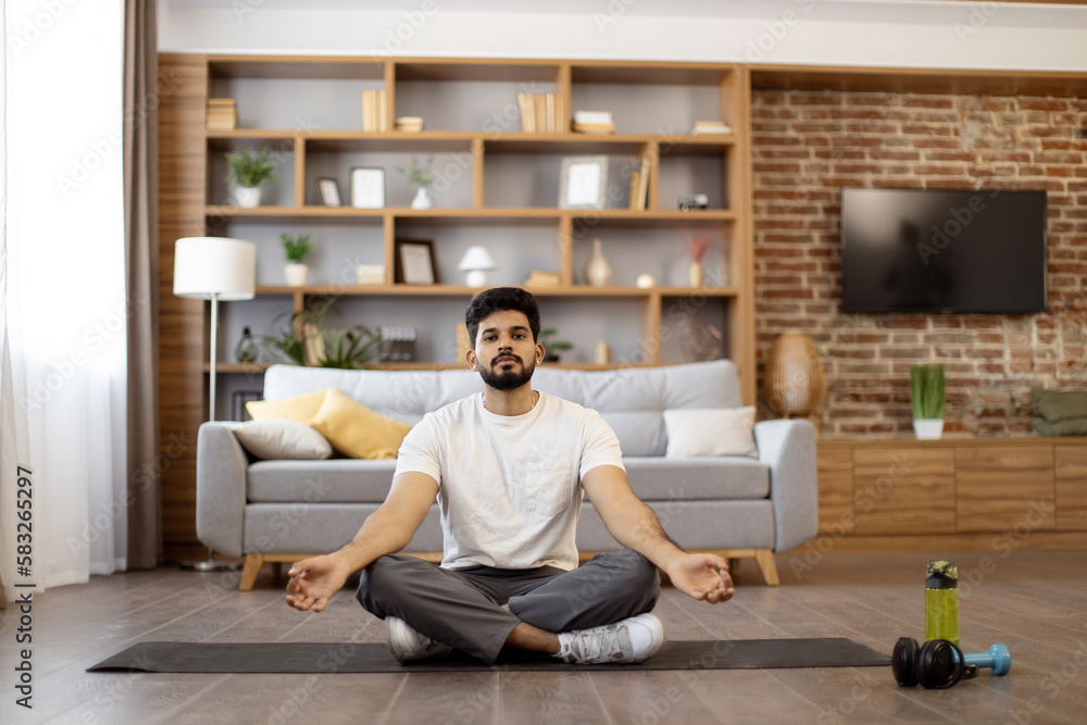 Relaxed indian man in sportswear practicing meditation in lotus pose at cozy living room. Young bearded guy keeping hands in mudra gesture and breathing calmly during workout. Relaxed indian man in sportswear practicing meditation in lotus pose at cozy living room. Young bearded guy keeping hands in mudra gesture and breathing calmly during workout.