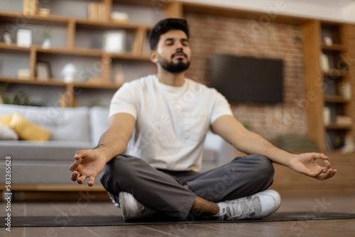 Handsome bearded arabian man in activewear keeping eyes closed and meditating on yoga mat at cozy living room. Concept of chakra, dzen and body wellness.