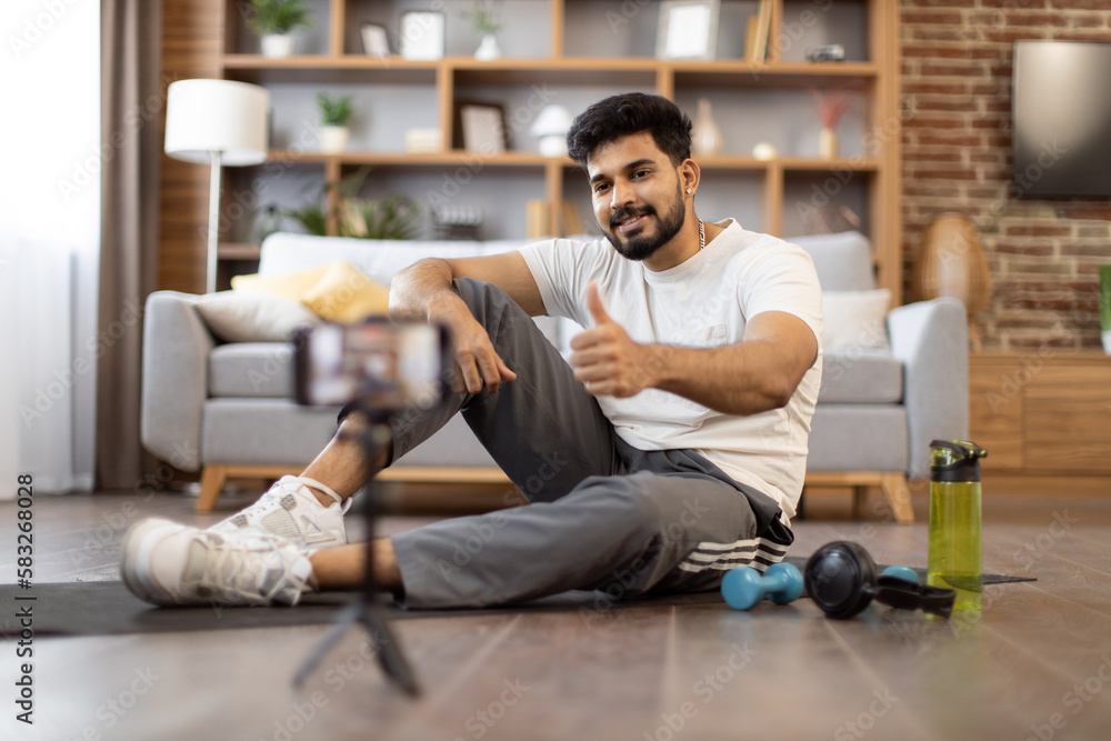 Young indian fitness expert sitting in front of phone on yoga mat near sports equipment for home workout showing thumb up. Active male blogger in gym wear recording home exercise for his online blog Young indian fitness expert sitting in front of phone on yoga mat near sports equipment for home workout showing thumb up. Active male blogger in gym wear recording home exercise for his online blog