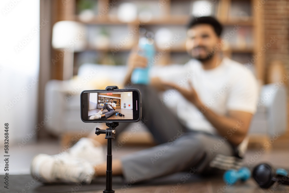 Focus on advanced mobile phone on tripod being used for making online tutorial about drinking water during home workout. Active arabic male pointing at sports bottle while sitting on floor indoors.
