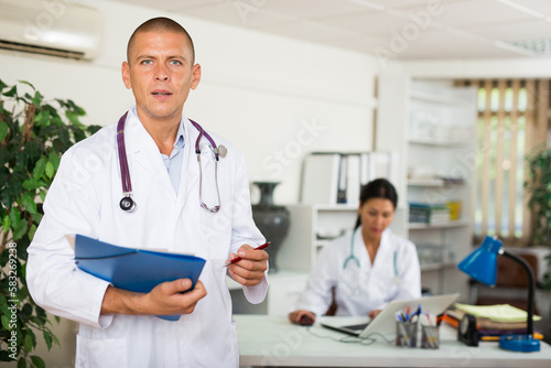 Portrait of polite doctor wearing white coat meeting patient in clinic office  filling out medical form at clipboard
