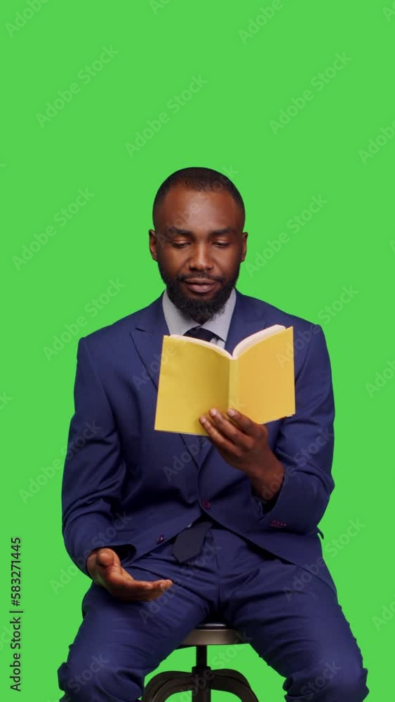 Vertical video: Front view of office worker reading literature book on camera, wearing business suit sitting on chair in studio. Confident person enjoying pages of novel or fiction story, posing over Vertical video: Front view of office worker reading literature book on camera, wearing business suit sitting on chair in studio. Confident person enjoying pages of novel or fiction story, posing over