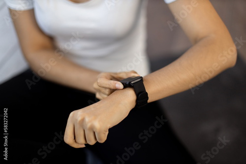 Close-up of young sporty woman resetting heart-rate watch on her wrist while sitting on yoga mat. Activity tracker for home training assisting fit athletic lady in meeting fitness objectives on time.