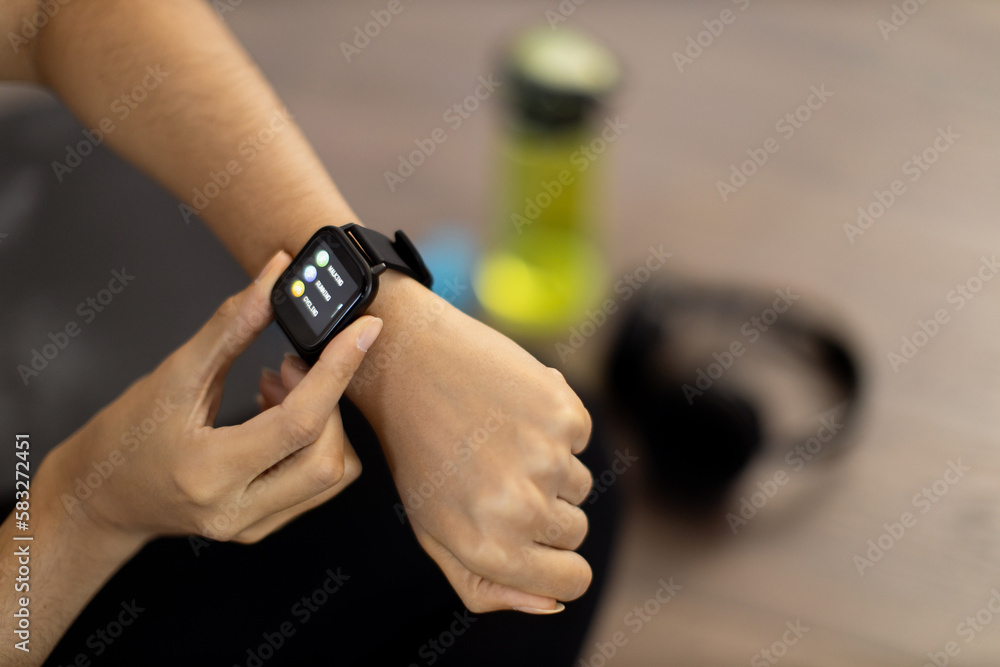 Close-up of young african american woman setting her modern smartwatch while sitting with bottle and headphones on floor. Active lady analysing valuable health and fitness data on activity tracker.