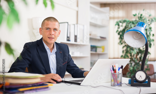 Positive businessman working with laptop and papers at office desk