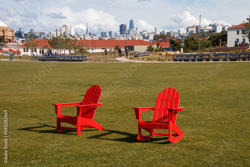 Two red chairs are sitting on green grass at The Presidio's Tunnel Top Park. In the background is the San Francisco cityscape. There is a blue sky with white clouds. photo