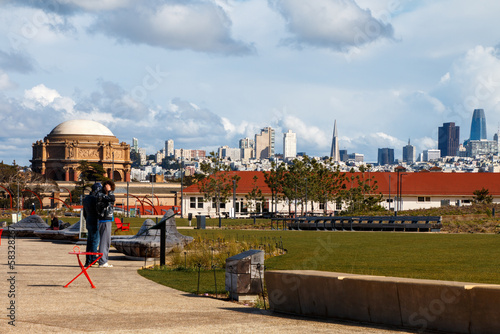 A view of the San Francisco skyline from the Tunnel Top park in The Presidio. Two people are standing to the side. There white clouds in a blue sky. photo
