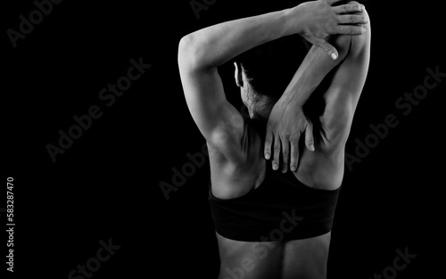 Serious female sporty muscular with ponytail doing stretching workout the shoulders  blades and neck in sport bra  holding hand behind the back on dark background. Back view.