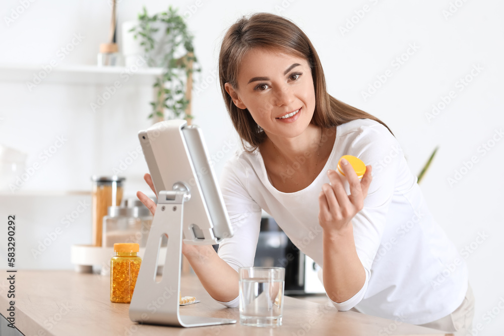 Beautiful woman with bottle of vitamin supplements and tablet computer in kitchen Beautiful woman with bottle of vitamin supplements and tablet computer in kitchen