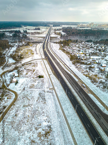 Top view of the roadway in winter. Snow-covered road and roadside. The highway passes through the forest and the private sector. Detailed photos from the drone of the road infrastructure.