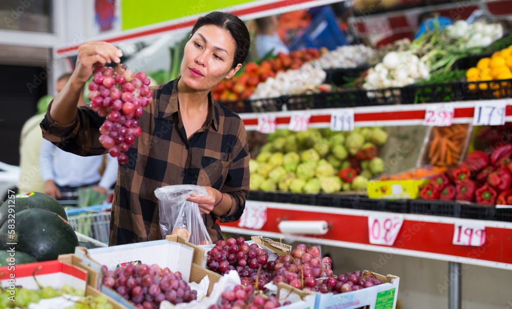 Oriental woman standing and choosing fresh ripe grape in greengrocer.