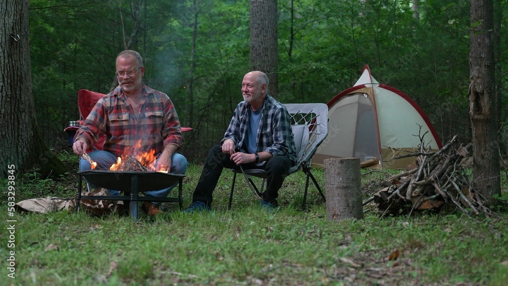 Two gay men building a campfire in a forest talking and laughing with tent in background.