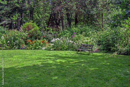 Fototapeta Naklejka Na Ścianę i Meble -  Spring. Resting place at Paulik Neighbourhood Park of Richmond City. Bench under the canopy of  tree on a green lawn with flower beds among flowering shrubs, British Columbia, Canada