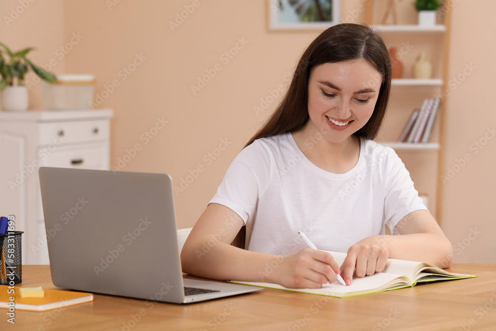 Woman writing in notebook while working on laptop at wooden table indoors Woman writing in notebook while working on laptop at wooden table indoors