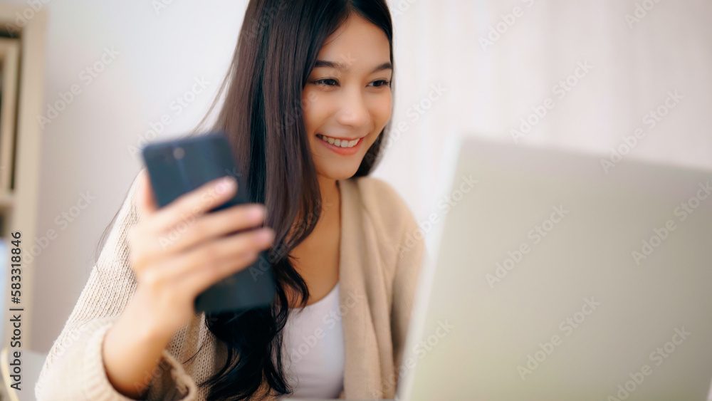 Young asian woman happy working on computer laptop and hand typing chat message on mobile smartphone at home. Video call, Online meeting Young asian woman happy working on computer laptop and hand typing chat message on mobile smartphone at home. Video call, Online meeting