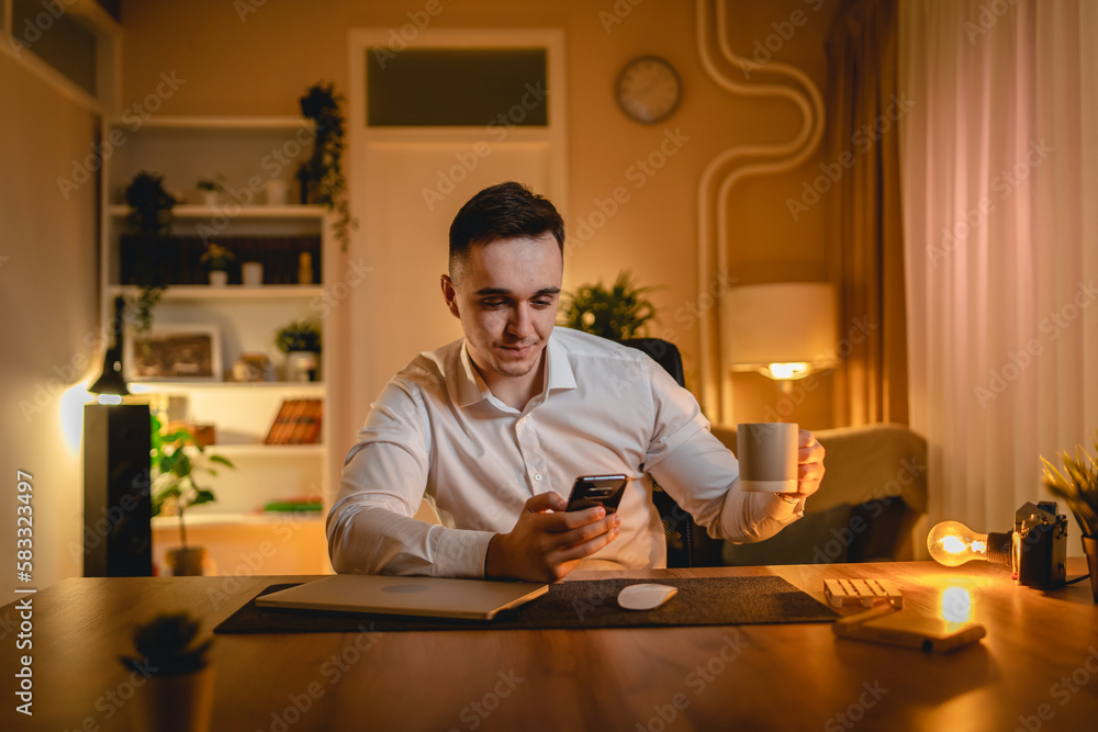 A young man is working late or studying at his home office while drinking coffee and using his mobile phone	