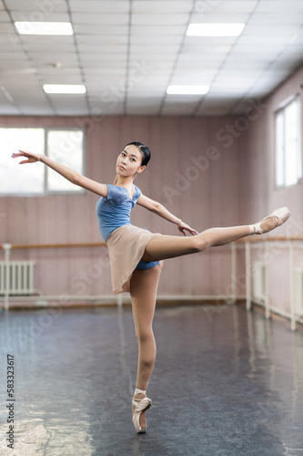 Graceful Asian ballerina in a blue bodysuit and a beige skirt is rehearsing in a dance class. 